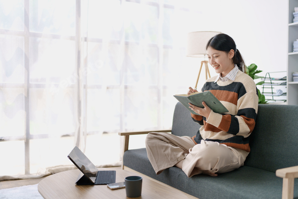A woman sits on a sofa, using her notebook and tablet computer simultaneously to study online, effic - Stock Photo - Images
