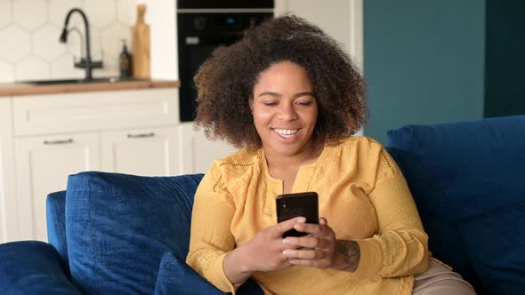 Happy AfricanAmerican Woman Using Smartphone Sitting on the Couch alt