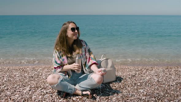 Young millennial woman drinking water from a metal bottle and sitting on the seashore on a sunny day alt