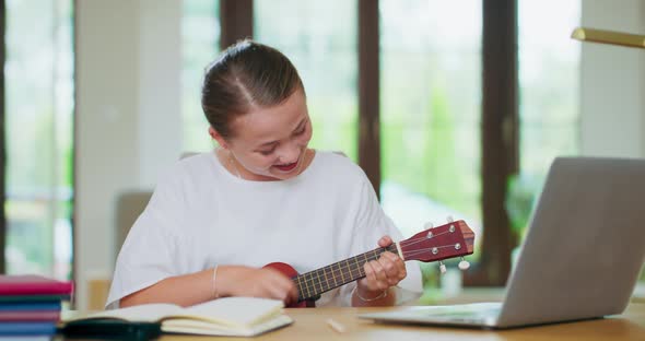 Teen Girl at the Desk in Front of Laptop Holds Smartphone and Writes Down Something Into Notebook alt