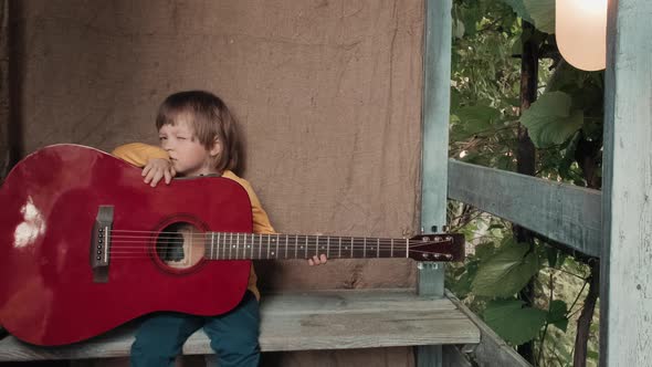Funny Child with Large Acoustic Guitar is Sitting on Porch of a Country House