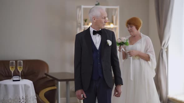 Worried Senior Caucasian Groom Checking Time on Watch Sighing As Blurred Bride Passing at Background alt