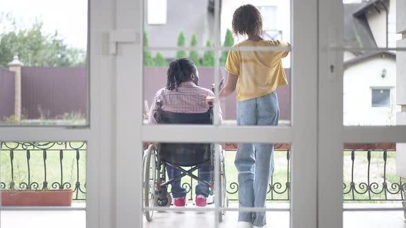 Back View Wide Shot Disabled Man in Wheelchair and Teenage Girl Standing on Porch Talking alt