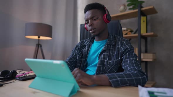 Young Man in Headphones and Pajamas Turning on Playlist on Tablet Sitting in Home Office Indoors alt