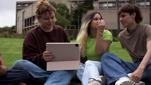 Young people studying outside of school building sitting on meadow and using digital tablet