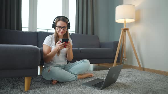 Casually Dressed Woman with Headphones Is Sitting on Carpet with Laptop and Smartphone and Working alt