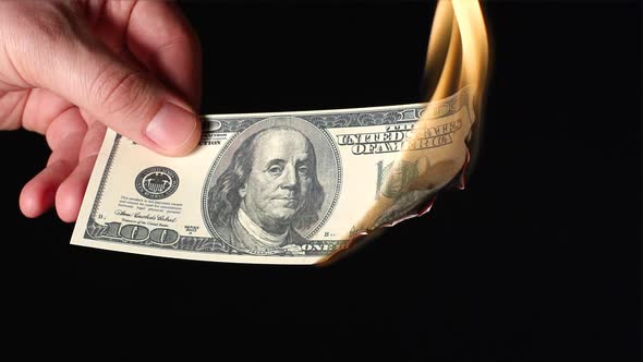 A human hand holds one hundred dollars flaming on a black background