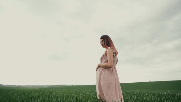 Young pregnant woman in a pink summer dress walks forward on a green field with wheat. 