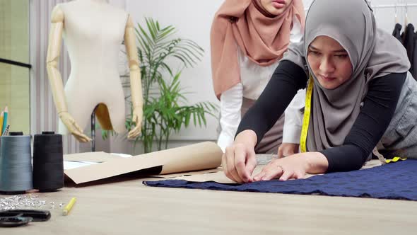 Muslim dressmaker pinning paper pattern on fabric at the table working with her assistant