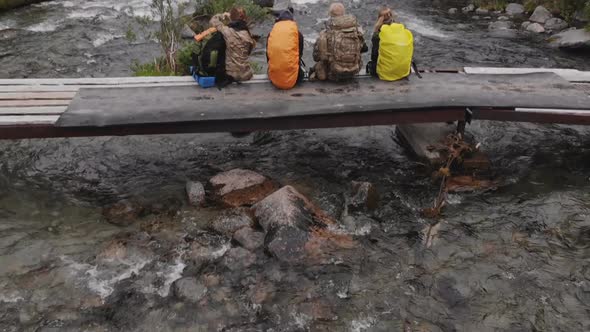Group of Friends or Tourists Sitting on Bridge in Mountains Area and Having Rest