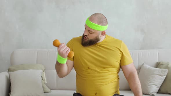 Funny Fat Man in Sportswear Lifting Dumbbells By Hands Sitting on Sofa at Home alt