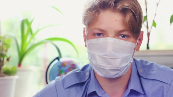  Teenager of European Appearance in a Blue Shirt Sits at Home, Puts on a Medical Protective Mask alt