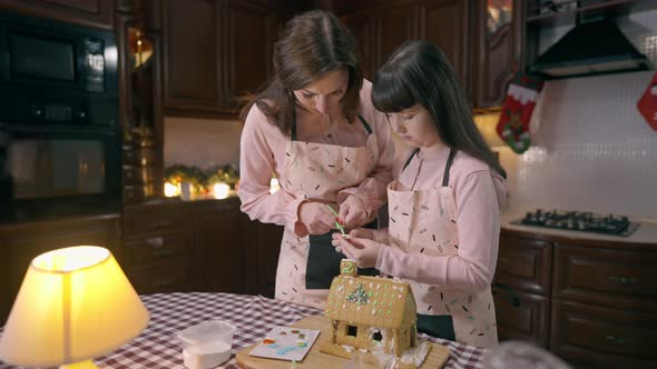 Charming Caucasian Girl Placing Santa on Decorated Gingerbread House Helping Woman Cooking Dessert alt