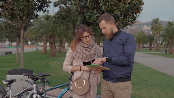 Young Man and Woman Are Viewing Map of City in Tablet Screen Standing in Park alt