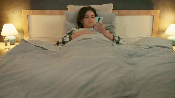 Brunette Girl Lying in Bed in the Evening and Reading Messages on Her Cell Phone Front View alt