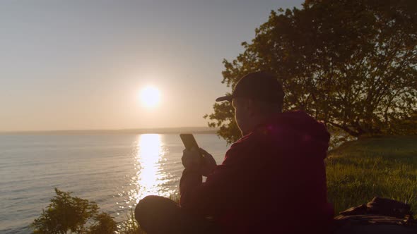 Man Tourist is Resting on the Seashore with a Smartphone in His Hand alt