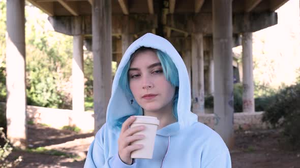 Blue haired teenage girl in oversize hoodie drinking coffee to go against bridge pillars alt