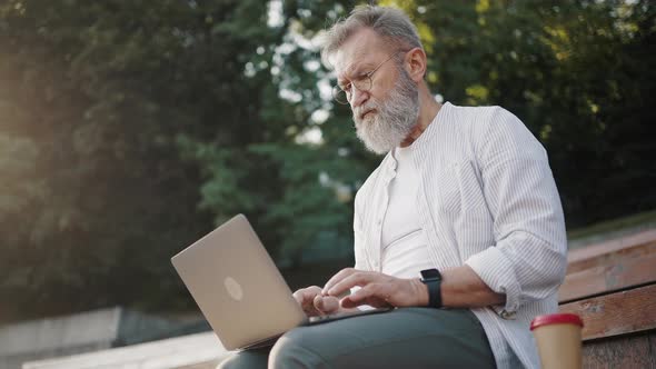 Bearded Man in Glasses Sits on Stairs Working on Laptop alt