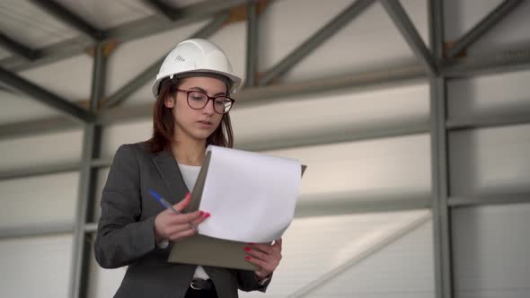 Young Woman in a Helmet with Documents at a Construction Site. The Boss Woman in a Suit Keeps alt