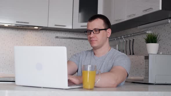 A Freelancer Man Works From Home at a Laptop While Sitting at a Table and Drinks Orange Juice alt