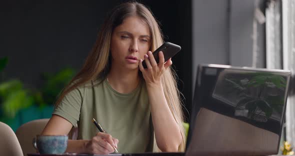 Woman Sitting in Cafe Holding Pen and Making Notes in Diarylaughingtalking on Speaker Using Phone alt