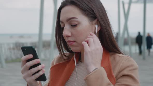 Attractive Young Woman Listening to Music in Headphone Use Smartphone at City Walk Look Around Smile alt