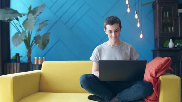 Woman with short hair is sitting on a yellow sofa with legs folded and typing on a laptop. freelance alt