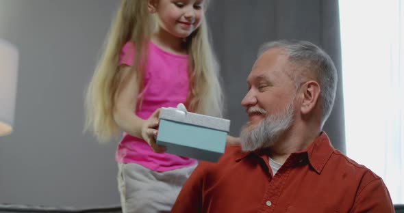 Cute Kid Daughter Make Surprise Present Covering Eyes of Happy Grandpa Receive Gift Box Sit on Sofa alt