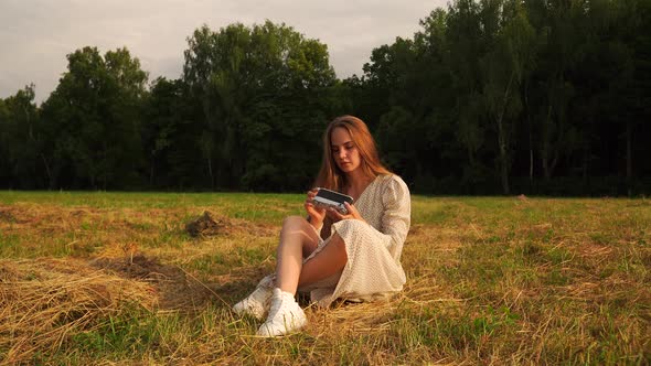 Beautiful Woman Sits the Ground in a Field Examines and Touches a Retro Camera