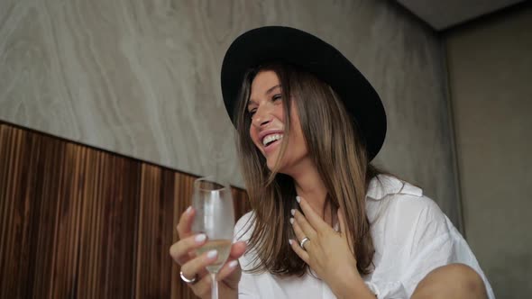Cheerful Girl in a Hat Laughs and Drinks Champagne Sitting on the Bed