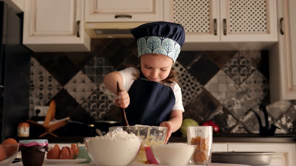 Little Girl in a Cook's Cap in the Kitchen, She Ineptly Mixes the Dough in Plate alt