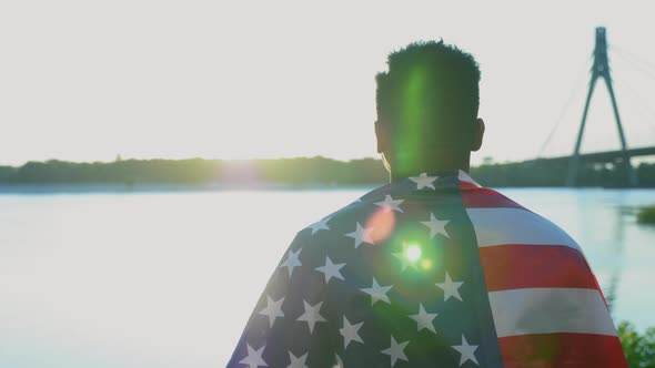 Afroamerican Man with American Flag on Shoulders Looks Into Distance at Sunrise alt