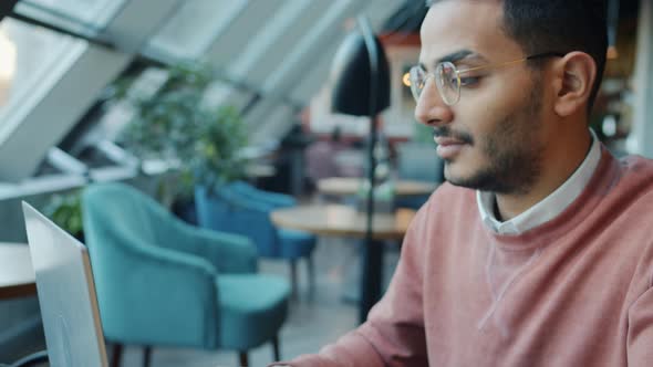 Tiltdown Portrait of Serious Arab Man Working with Laptop Sitting at Table in Modern Cafe Typing alt