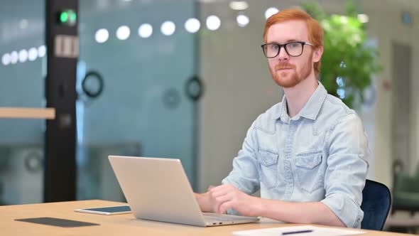 Thumbs Up By Young Redhead Man Working on Laptop in Office alt