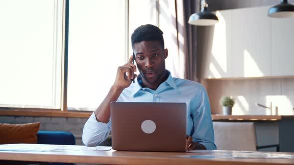 Angry young man is talking on mobile phone and working with laptop in the kitchen alt