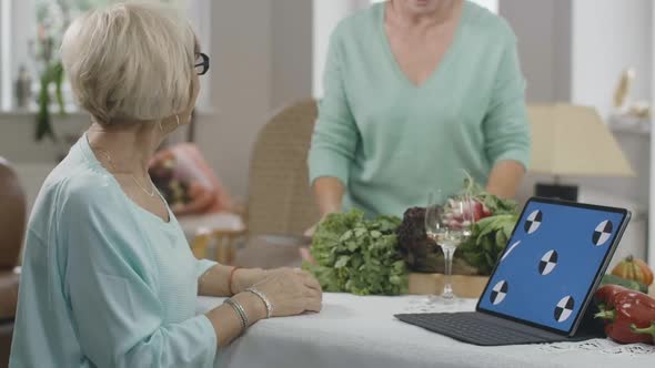 Side View of Slim Senior Woman Reading Recipe From Webpage on Blue Screen Laptop As Unrecognizable alt