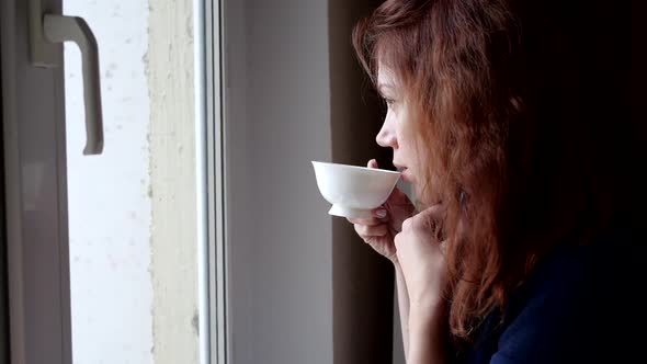 Young Woman Drinks Coffee in Medical Mask Looking Out the Window During Quarantine. Social