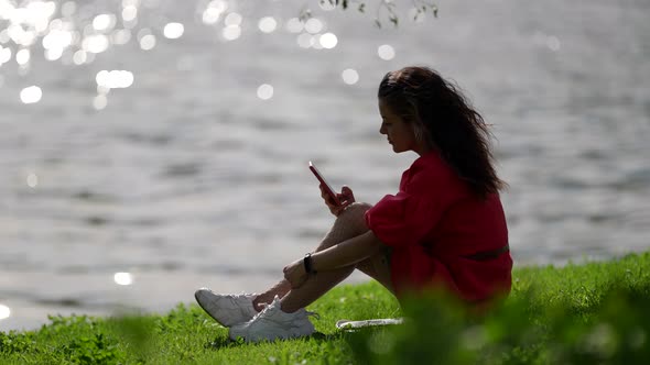 Relaxed Young Woman is Spending Time on Shore of River or Lake and Using Smartphone alt