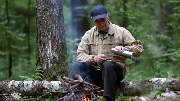 an Elderly Man in a Cap is Sitting in the Forest on a Log in Front of a Fire and Pours a Drink From alt