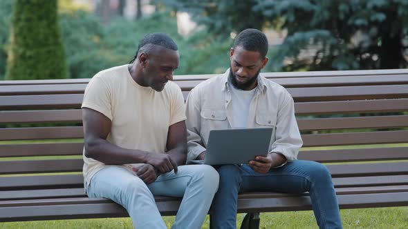 Two Black Men in Casual Clothes Sitting on Bench in City Park Using Laptop alt