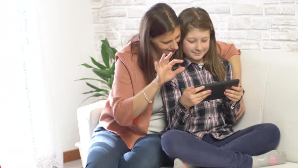 Happy mother and her daughter smiling having fun using digital tablet sitting on white sofa at home alt