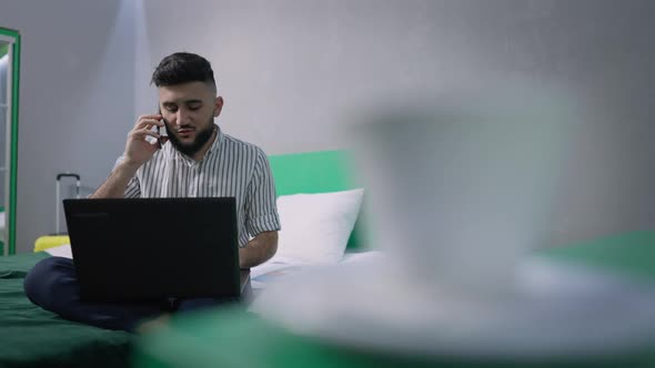 Confident Young Businessman Talking on the Phone Sitting on Bed in Hotel Room with Blurred Tea alt