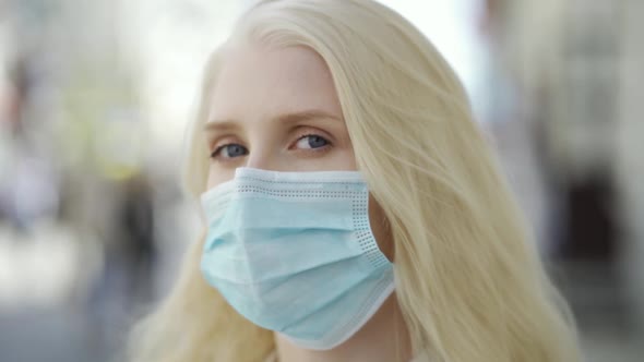 A Blueeyed Blonde Woman in a Medical Mask Stands on the Central Street of a Big City and Looks Into