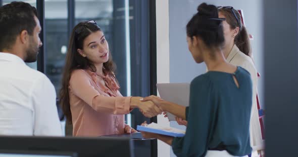 Two diverse businesswomen standing shaking hands during meeting with colleagues alt