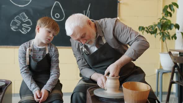 Loving Grandfather Making Ceramic Pot on Throwing Wheel and Talking To Grandchild alt