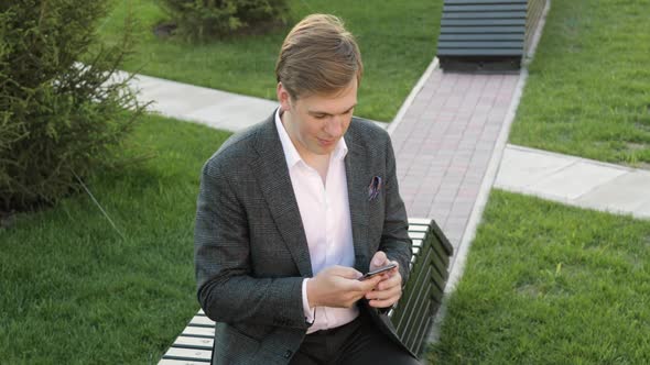 Businessman Is Typing on Smartphone Browsing Online Sitting on Bench in Park