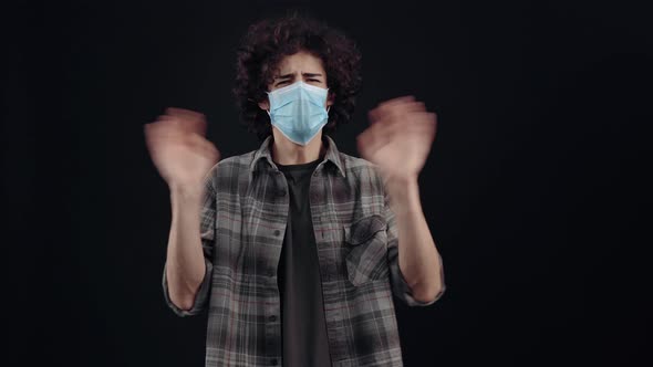 The Young Man with Curly Hair Wears a Mask Looks to One Side and Waves His Hands in a Sign That the alt