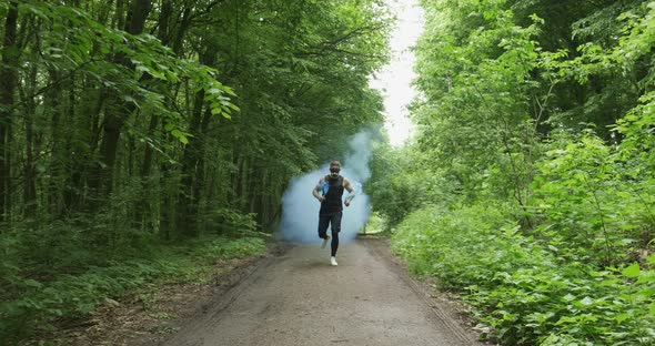Young Male with Face Mask in Sportswear Running with Blues Smoke Bomb on the Road at Forest. Men alt