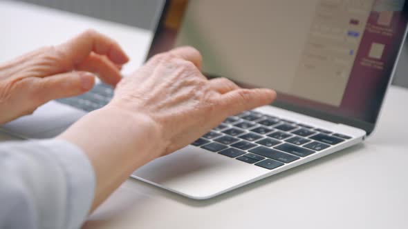 Old woman hands type on black keyboard of grey laptop on white table alt