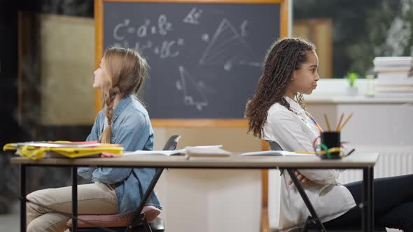 Argued Schoolgirls Sitting Back to Back in Classroom Talking and Showing Tongue Out alt